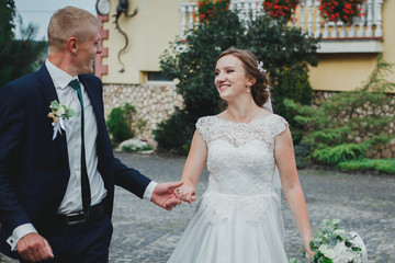 Beautiful blonde couple is running outside near the old stone house walls on the cobblestone road. Smiling groom with bride in satin lace dress are holding hands and having fun.