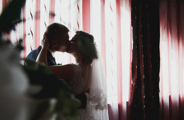 Beautiful wedding couple is kissing near the window. Bride and groom silhouettes on the dark red background. Romantic meeting in the wedding morning. Black and white.