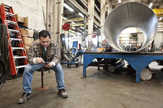 Hispanic man factory worker checking a part inventory on his cell phone in a sheet metal factory.