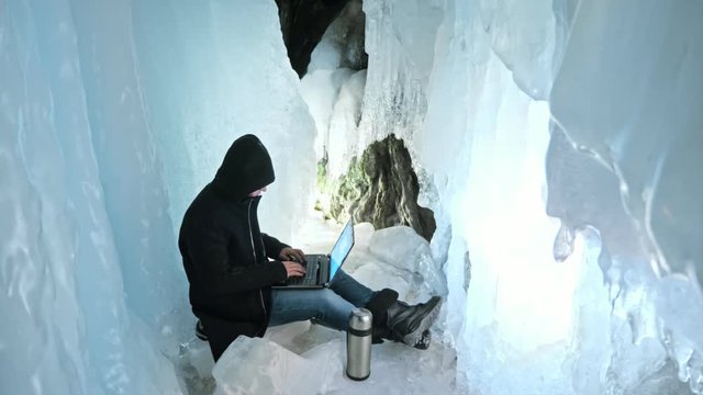 Hacker hacks the server. Man to program on laptop in ice cave. Around the mysterious ice grotto. Man hides himself in a hood. Hacker has criminal cases on the Internet. Guy is warming himself with the