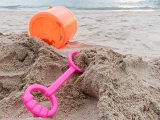 Fototapeta na wymiar Pink spade and orange plastic bucket of sand tool toy set for kid and toddler on the sand over beach, sea, and sky background