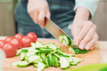 close-up of female hands cut into fresh cut cucumbers on a wooden cutting board next to pink tomatoes. The concept of homemade vegetarian cuisine and healthy eating and lifestyle