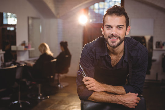 Smiling male hairdresser leaning on chair in salon