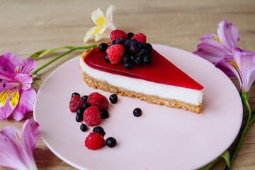 close-up of a pink plate with a cheesecake with jelly and berries around which lie the flowers of a wild orchid on a wooden table