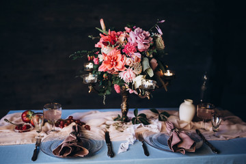 close-up decorated festive table with blue tablecloth vintage glasses, crockery and appliances and a floral bouquet with candles on the background of a wooden wall