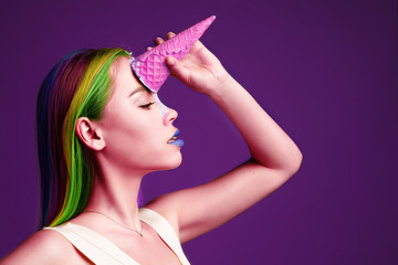 close-up of a girl in a pink dress holds and with multi-colored hair holds her hand near her forehead a pink waffle glass with ice cream as a unicorn on a purple background