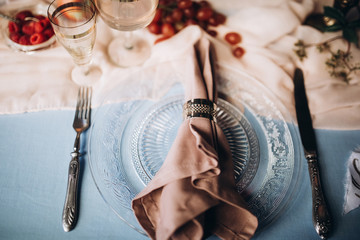 close-up of a napkin with a gold ring on a glass plate with vintage instruments on a decorated festive table with a blue tablecloth vintage glasses and flowers with candles on a wooden wall background