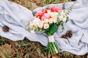 a wedding bouquet of pink roses and peonies that lays on a gray cloth with cones on the ground in the forest