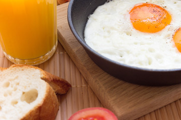 Breakfast with two fried eggs in pan, slice of french bread, tomato, fresh orange juice on mat