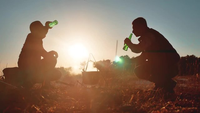 two tourists beer are sitting lifestyle by bonfire party in nature camping silhouette sunlight sunset. two men silhouette on sunlight vacation cauldron lights fire the bonnet the sausages and drink