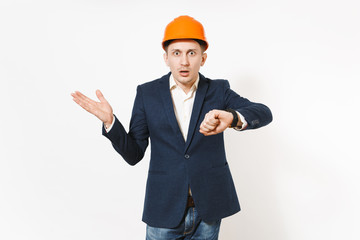 Shocked businessman in dark suit, protective construction orange helmet with clock spreading hands isolated on white background. Time is running out. Male worker for advertisement. Business concept.