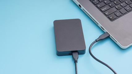 Black external hard disk connecting to a laptop on a blue background. - obrazy, fototapety, plakaty