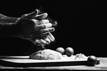 Beautiful and strong men's hands knead the dough from which they will then make bread, pasta or pizza. A cloud of flour flies around like dust. Next to the chicken egg. The background is dark.