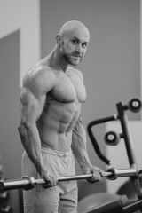 Fototapeta na wymiar Shredded shirtless bodybuilder with beard and shaved head doing biceps curls with barbell in a gym. Fit man trains. Black and white portrait.