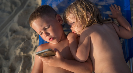 Little caucasian brother and sister looking at cell fhone lying at sandy beach