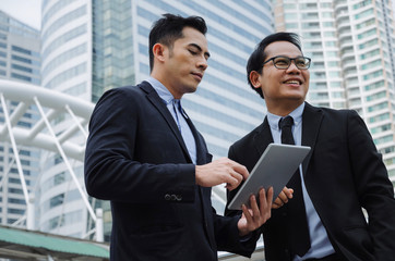 Naklejka na ściany i meble two business young asian smart man talking and reading information about finance news in mobile tablet together standing in modern city, network technology, internet, successful, teamwork concept