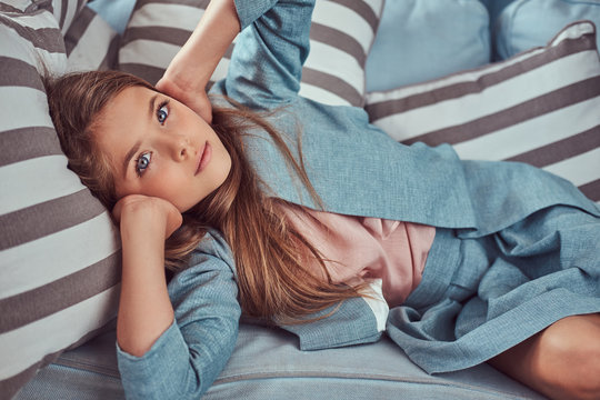 Portrait of a cute little girl with long brown hair and piercing glance, looking at a camera, lying on a sofa at home alone