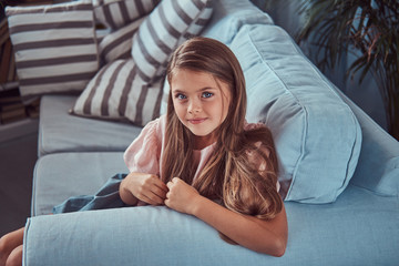 Portrait of a smiling little girl with long brown hair and piercing glance, lying on a sofa at home