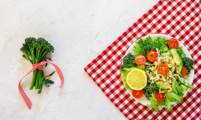 Broccolini vegan salad with lettuce, avocado, tomatoes, sprouts, lemon on white plate on white squares napkin 
on grey concret background, top view, copy space, Healthy food and diet concept