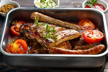 Grilled pork ribs and sausages served with tomato and sauces on wooden table.