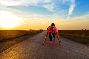 Athletic woman in a start position ready to run on rural road during sunset.	