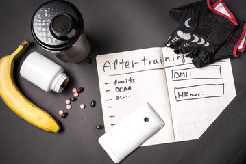 Top view of fitness table, gym equipment, training diary, protein shaker, vitamins and banana on dark background