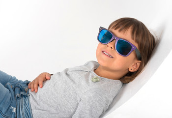 A little kid in glasses is lying and smiling in the studio against the white background.