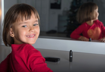 The little girl in a red jacket smiles in front of the mirror in a photographic studio.