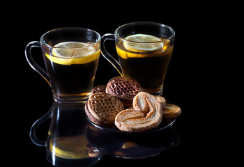 Black tea with a lemon, with cookies in a glass cup, with reflection on a black background.