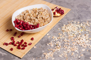 Cooked yummy warm oatmeal and heap of ripe fresh pomegranate seeds in white ceramic bowl near piece of pomegranate on clean bamboo rug beside scattered dry oat on worn gray scratched concrete