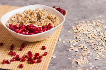 Cooked yummy warm oatmeal and heap of ripe fresh pomegranate seeds in white ceramic bowl near piece of pomegranate on clean bamboo rug beside scattered dry oat on worn gray scratched concrete