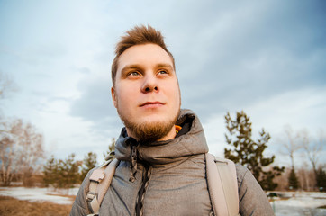 Ginger young guy with beard looks up against the sky, trees and clouds.