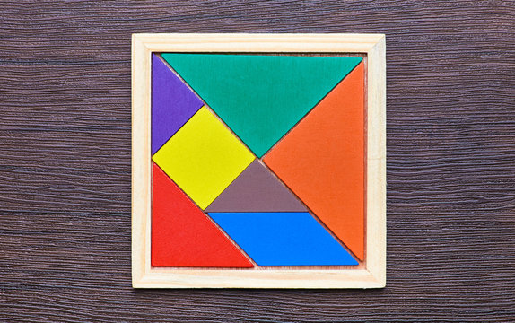A tangram puzzle consisting of colored pieces with geometric shapes, collected in a square on a dark wooden background