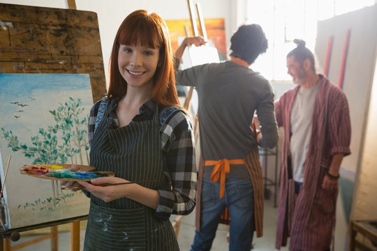 Portrait of woman holding color palette while friends painting in background