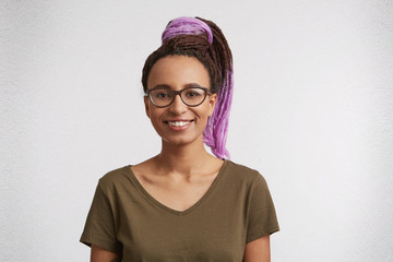 Indoor portrait of beautiful mixed race woman with shaggy hairstyle smiling cheerfully, showing her white teeth to the world. Wears glasses and t-shirt.