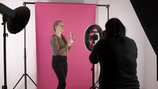 Stock photographer taking pictures in studio. Young model as vlogger recording video blog. Backstage shot during photoshoot