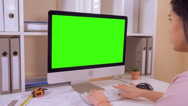 back view woman with black long hair sitting at the workplace working on computer. architect sitting at the white desk with building plan in front of display with isolated green screen