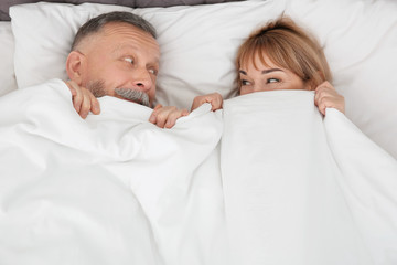 Mature couple hiding together under blanket in bed at home