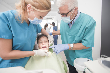 Boy with perfect teeth at the dentist doing check up with two doctors while other doctors analyze x-ray in the background - oral hygiene health care concept