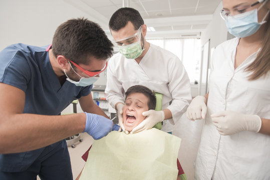 Scared teenager boy is having a check up at the dentist while being held byu three doctors
