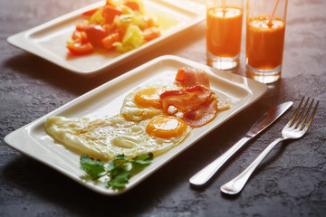 Breakfast from fried egg, bacon, vegetable salad and carrot juice. On a dark stone background