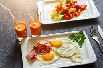 Breakfast from fried egg, bacon, vegetable salad and carrot juice. On a dark stone background