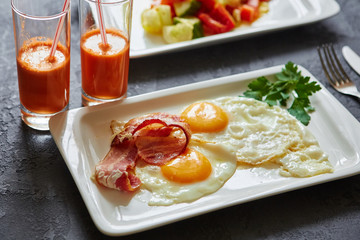 Breakfast from fried egg, bacon, vegetable salad and carrot juice. On a dark stone background