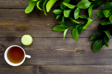 Summer mood, summer tea party. Cup of tea and sweets macarons near brignt greenery on dark wooden background top view copy space