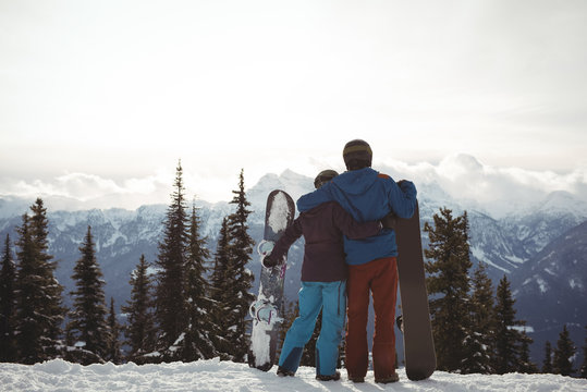 Rear view of couple holding snowboard at mountain during winter against sky