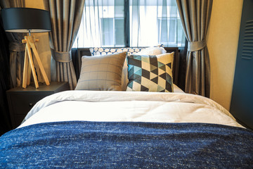 Many pillow and a book with white and brown blanket on the bed with brown wooden lamp side bed on the table.