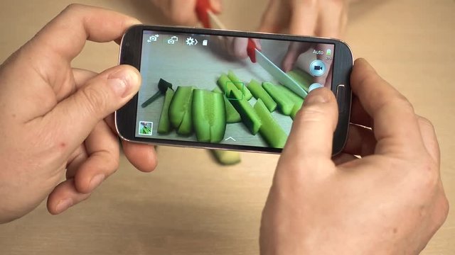 A man takes pictures as a woman cuts cucumbers with a knife. The husband takes pictures on his smartphone as his wife prepares a dish.