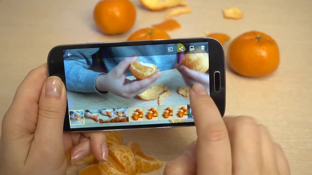 Woman's hand, mother takes pictures on a smartphone, as a child cleans, peeling and eats a mandarin. A girl 5-6 years old is sitting at a children's table.