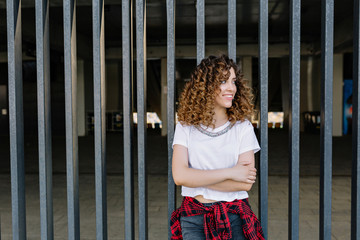 image of fantastic girl with open smile and curly style hair dressed in white shirt and checkered shirt in the city. She is looking aside. Real happy emotions. Place for text.