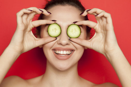 Happy young woman posing with slices of cucumber on her face on red background